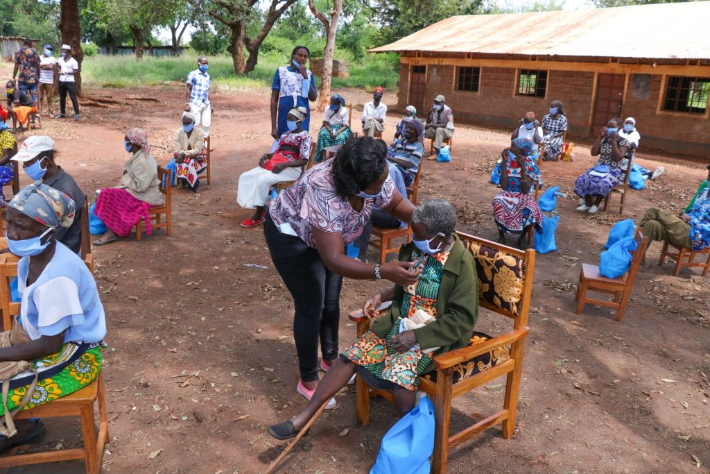 High angle of anonymous African female taking care of senior people in face masks sitting on chair near shabby building of nursing home in village